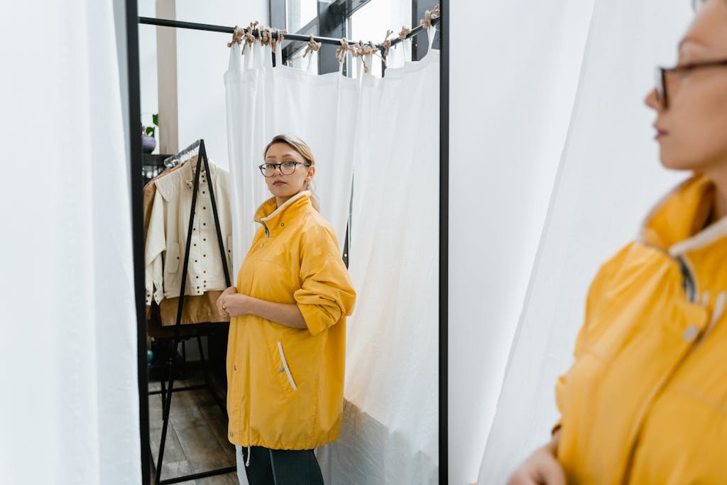 A woman in a yellow coat examines her reflection in a fitting room mirror surrounded by trendy clothing.