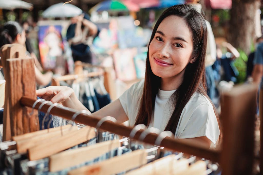 Glad Asian female standing near track with clothes and choosing garments while spending sunny day in street market
